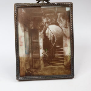 May include: A rectangular metal picture frame with a textured border and a small bow-shaped ornament at the top. The frame contains a sepia-toned image of an interior scene featuring a wooden door, a curved staircase, and a small green plant.