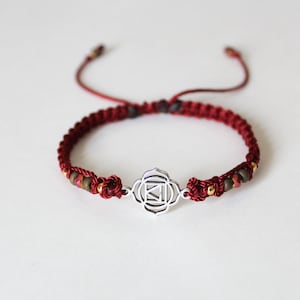 May include: Red string bracelet with a silver charm featuring the symbol for the sacral chakra. The bracelet is accented with small brown, gold, and red beads.