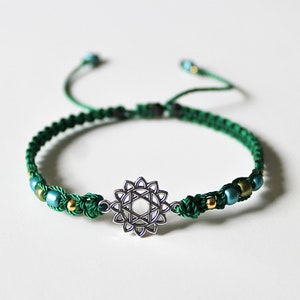 May include: Green braided bracelet with a silver charm featuring a geometric design. The bracelet is accented with small green and gold beads.
