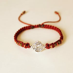 May include: A red cord bracelet featuring a central silver-toned metal charm with a geometric symbol. The band is constructed from braided red thread and includes small metallic and brown beads integrated into the design with adjustable pull strings.