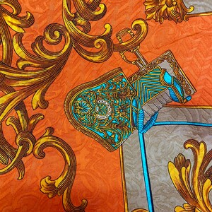 K&ouml;nnte beinhalten: Nahaufnahme eines Stoffes mit einem sich wiederholenden Muster aus goldenen Schn&ouml;rkeln und einem blauen und goldenen Helmdesign. Der Hintergrund ist in einem warmen Orangeton gehalten.