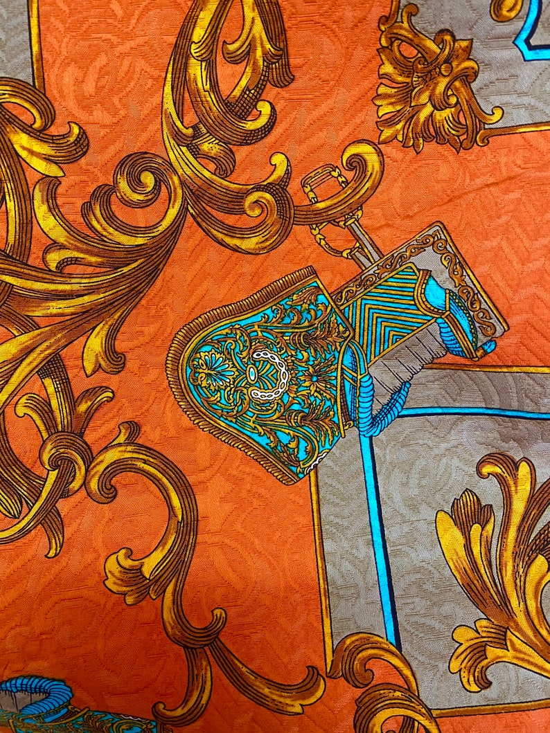 K&ouml;nnte beinhalten: Nahaufnahme eines Stoffes mit einem sich wiederholenden Muster aus goldenen Schn&ouml;rkeln und einem blauen und goldenen Helmdesign. Der Hintergrund ist in einem warmen Orangeton gehalten.