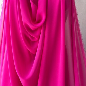 Op de afbeelding: Een helder roze, doorschijnende stof met een zachte, vloeiende textuur. De stof is gedrapeerd en gevouwen, waardoor de drape en transparantie worden getoond.