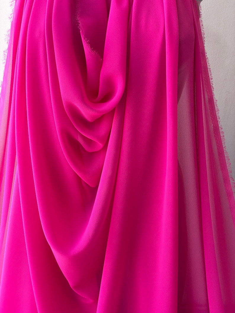 Op de afbeelding: Een helder roze, doorschijnende stof met een zachte, vloeiende textuur. De stof is gedrapeerd en gevouwen, waardoor de drape en transparantie worden getoond.
