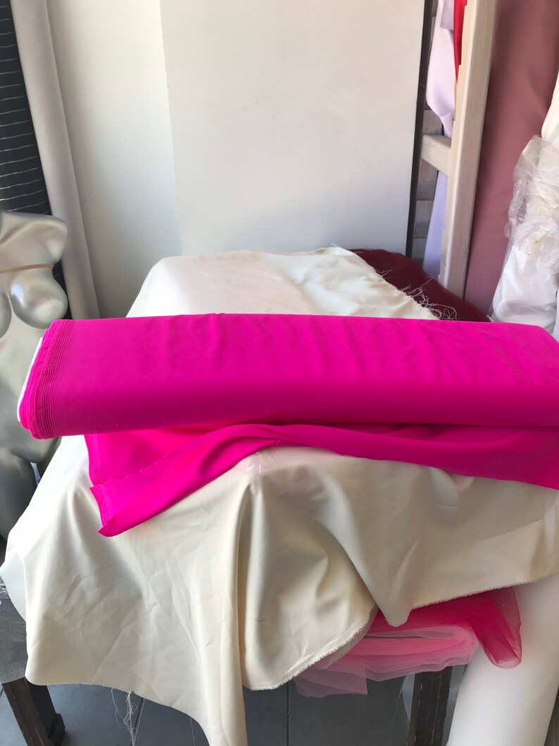 May include: A roll of bright pink fabric folded on top of a white fabric. The fabric is on a table with a white surface.