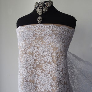 May include: White floral lace fabric draped over a mannequin. The lace features a delicate, repeating pattern of flowers. A silver skull necklace adorns the mannequin's neck, adding a touch of gothic flair. The fabric is displayed against a neutral background.