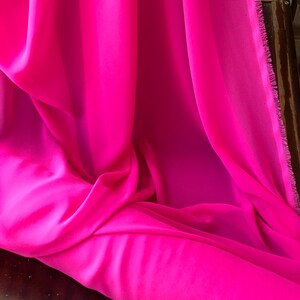 Op de afbeelding: Een helder roze, doorschijnende stof, gevouwen en gedrapeerd op een houten oppervlak. De stof heeft een zachte, vloeiende textuur.