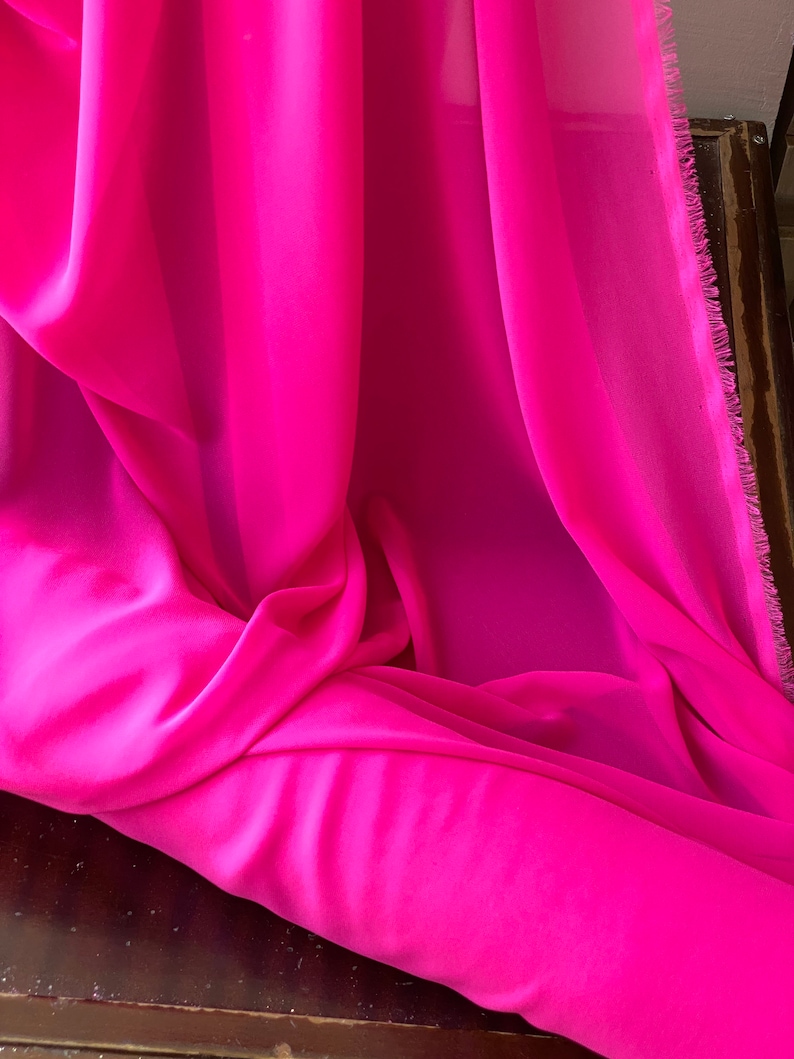 Op de afbeelding: Een helder roze, doorschijnende stof, gevouwen en gedrapeerd op een houten oppervlak. De stof heeft een zachte, vloeiende textuur.