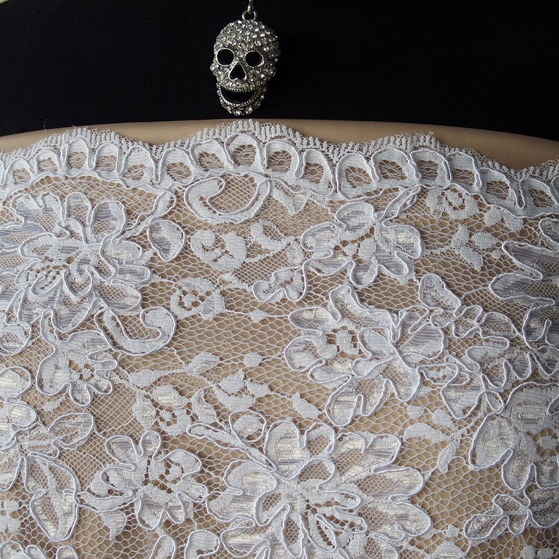 May include: A silver skull charm, embellished with crystals, is suspended above a white floral lace. The lace displays an intricate pattern of flowers and leaves, set against a beige backdrop. The design is delicate and detailed, ideal for accessories or clothing.