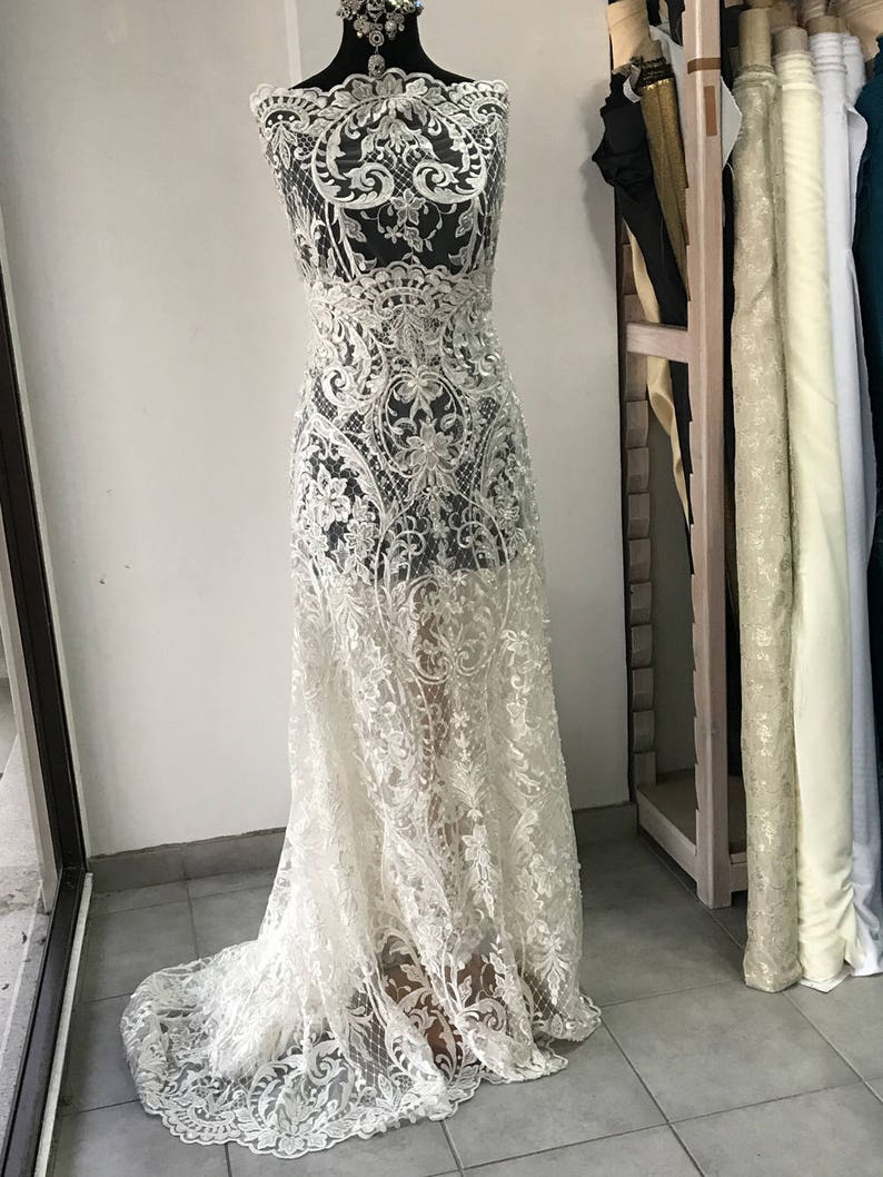 May include: White lace fabric with a delicate floral pattern. The fabric is displayed on a dress form and has a flowing, elegant design.