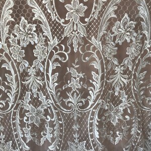 May include: White lace fabric with a delicate floral pattern and small sequins. The lace has a scalloped edge and is perfect for adding a touch of elegance to any project.