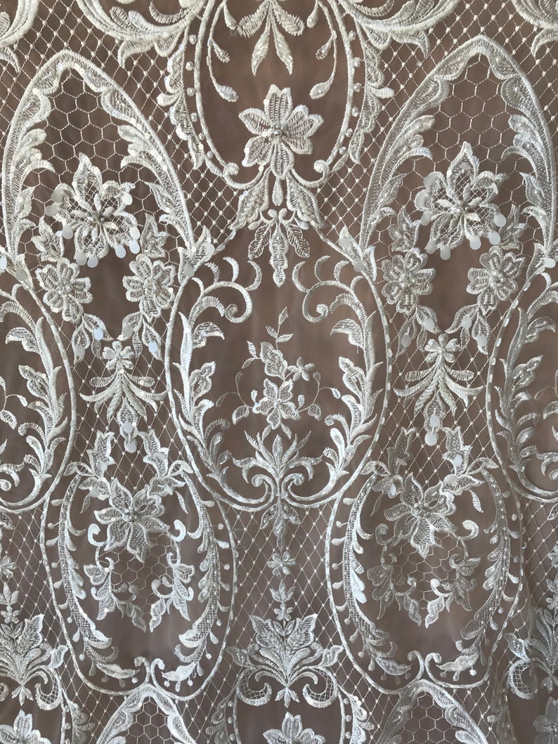 May include: White lace fabric with a delicate floral pattern and small sequins. The lace has a scalloped edge and is perfect for adding a touch of elegance to any project.