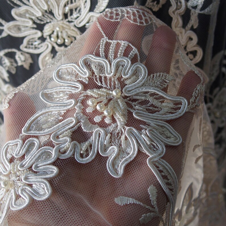 May include: Close-up of ivory lace fabric with a floral design and pearl embellishments. The lace is delicate and intricate, with a soft, flowing texture.