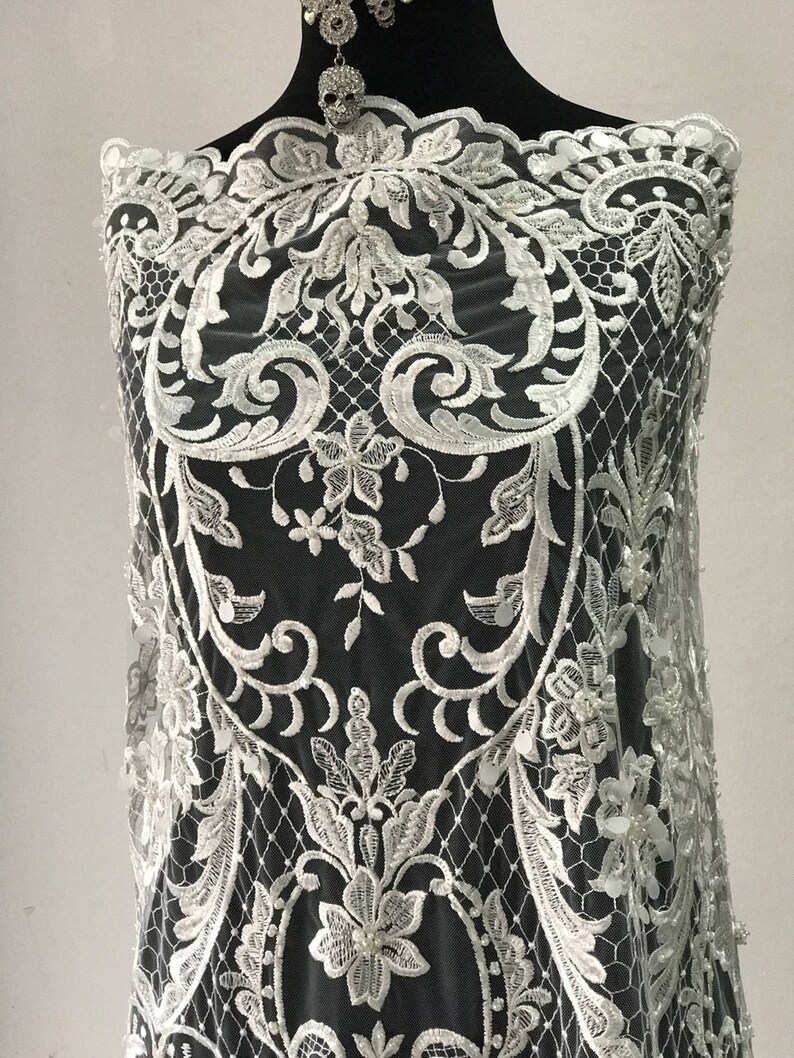 May include: White lace fabric with a floral pattern and pearl embellishments. The lace is delicate and intricate, and the pearls add a touch of elegance.