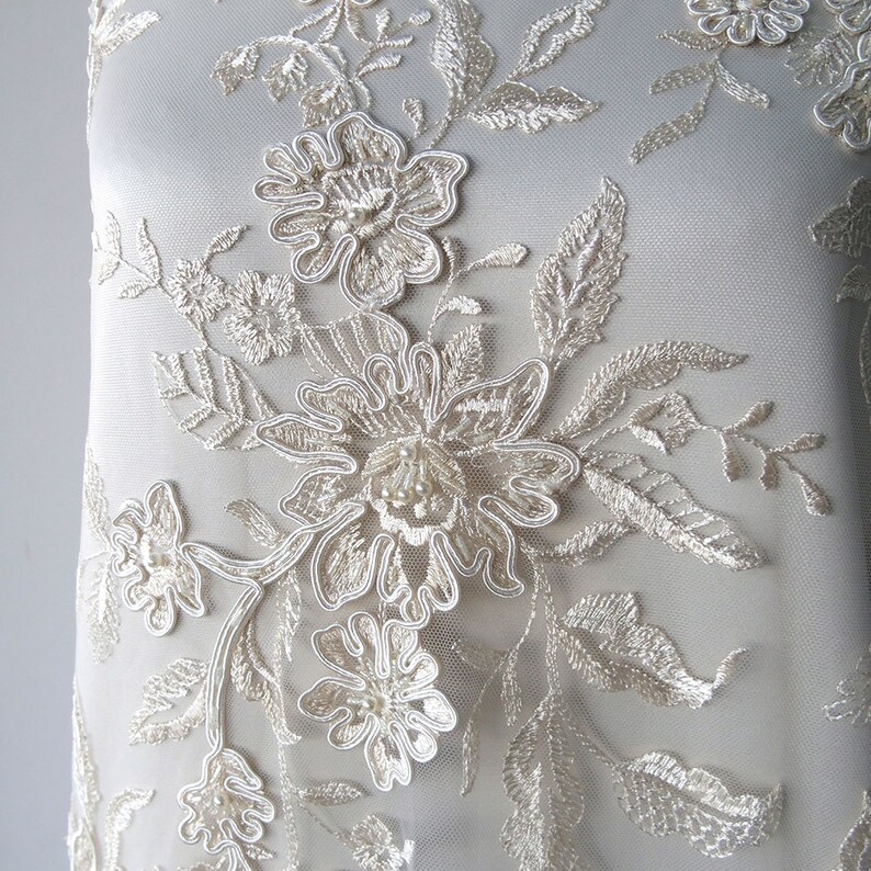 May include: Ivory lace fabric with a floral design. The flowers are 3D and have a beaded center. The fabric is sheer and delicate.