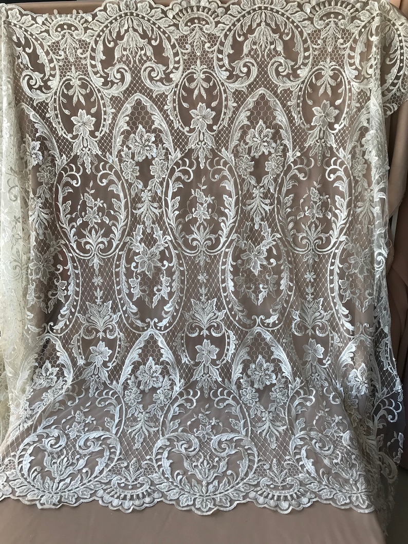 May include: White lace fabric with a delicate floral and scroll pattern. The lace has a scalloped edge.