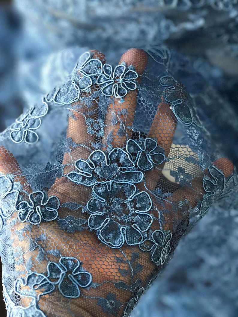 Op de afbeelding: Close-up van een lichtblauwe kantstof met een bloemenpatroon. De kant heeft een delicaat, ingewikkeld ontwerp met kleine bloemen.