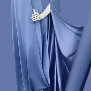 Può includere: Un manichino drappeggiato in tessuto blu pervinca fluente. Il tessuto ha una consistenza liscia, simile al raso, ed è disposto per suggerire un capo. Un rotolo dello stesso tessuto è visibile sullo sfondo.