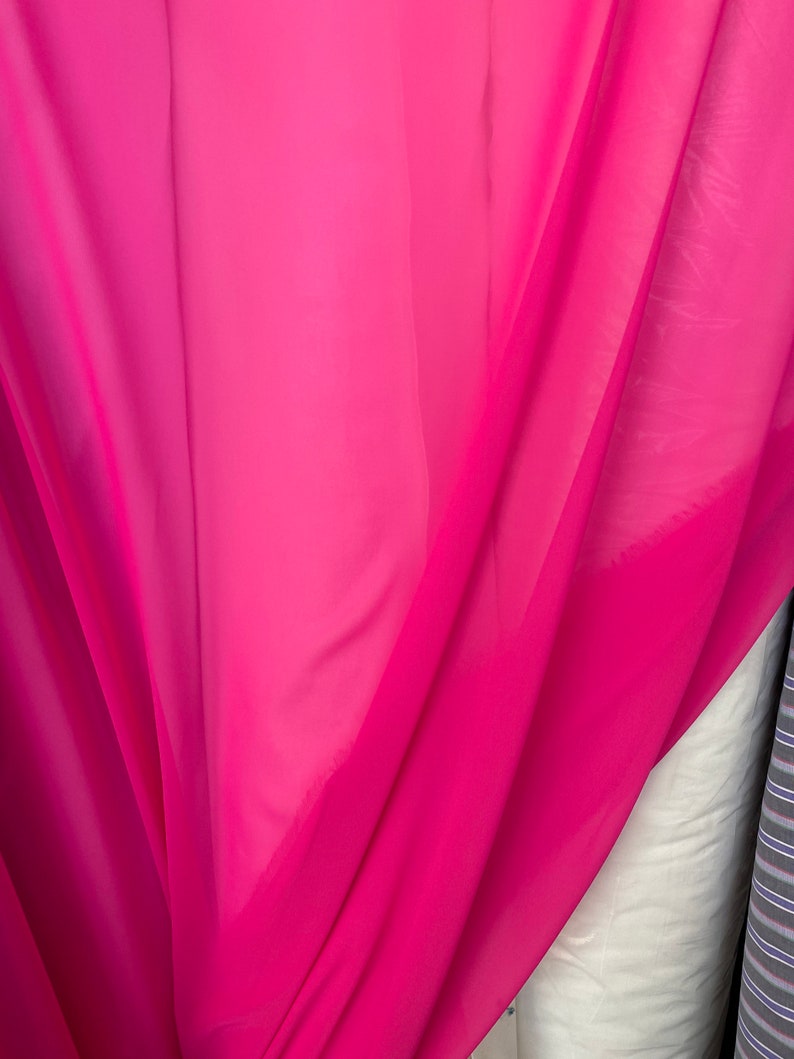 Op de afbeelding: Een helder roze, doorschijnende stof met een gladde textuur. De stof is gevouwen en gedrapeerd, wat de vloeiende aard laat zien.