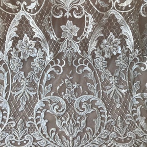 May include: White lace fabric with a floral pattern and sequins. The lace is delicate and intricate, with a scalloped edge.