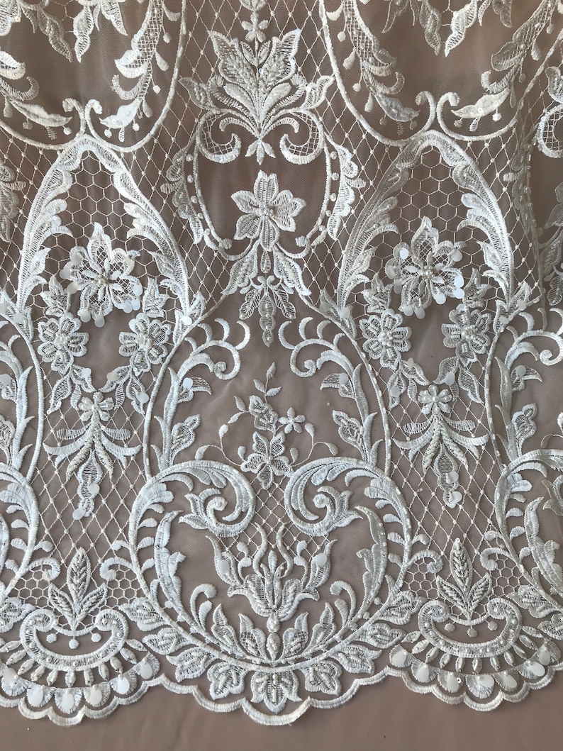 May include: White lace fabric with a floral pattern and sequins. The lace is delicate and intricate, with a scalloped edge.