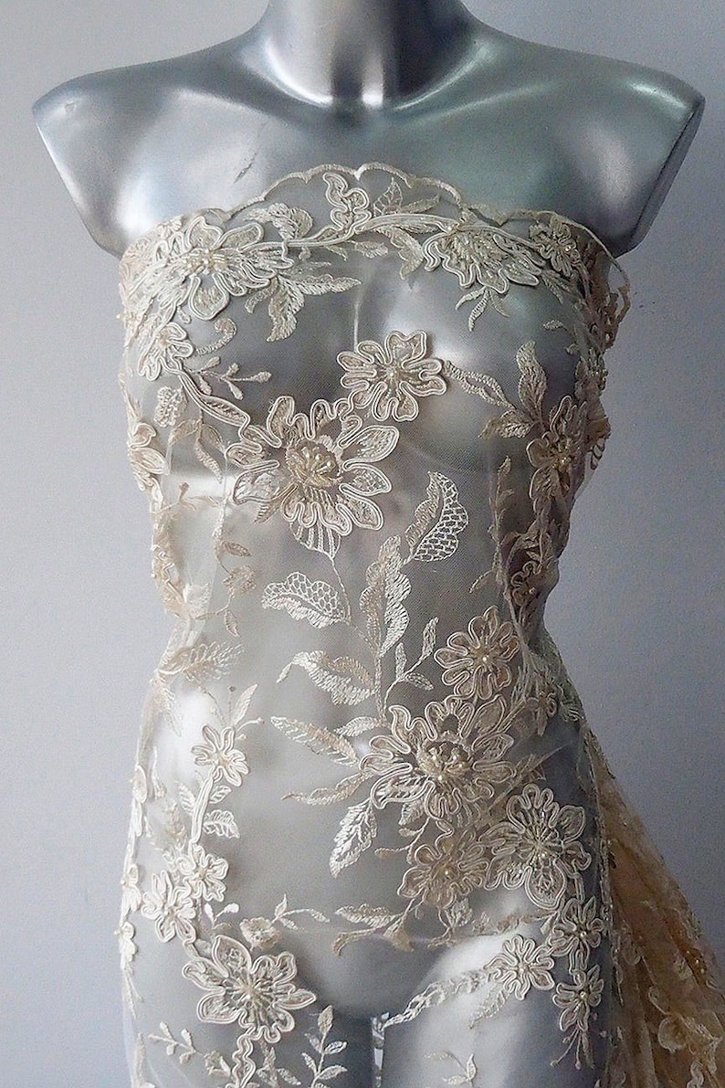 May include: Ivory floral lace fabric with pearl embellishments. The lace features a delicate floral pattern with intricate details and a sheer mesh background.