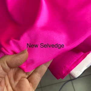 May include: Close-up of a bright pink fabric with a selvedge edge. The fabric is smooth and shiny, and the selvedge edge is visible. The text "New Selvedge" is printed on the fabric.