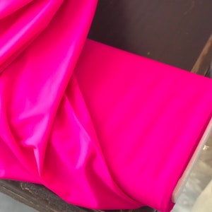 May include: A bright pink fabric folded on a wooden surface. The fabric is smooth and has a slight sheen.