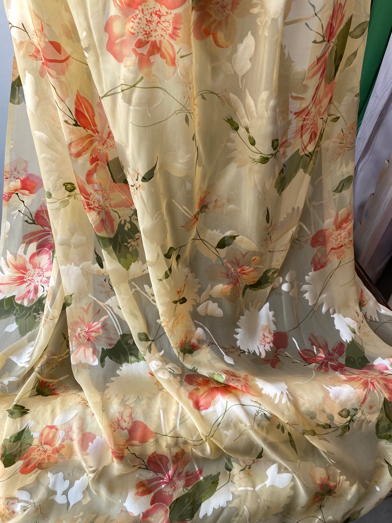 May include: Sheer, light yellow fabric with a floral print of pink and orange flowers. The fabric is draped and has a soft, flowing texture.
