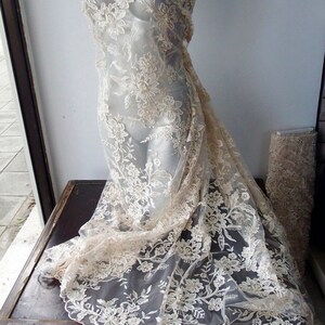 May include: A delicate ivory lace fabric with a floral pattern. The lace is draped over a mannequin and has a soft, flowing texture.