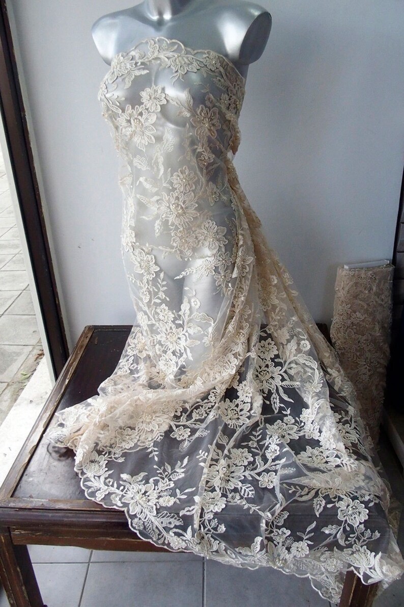 May include: A delicate ivory lace fabric with a floral pattern. The lace is draped over a mannequin and has a soft, flowing texture.