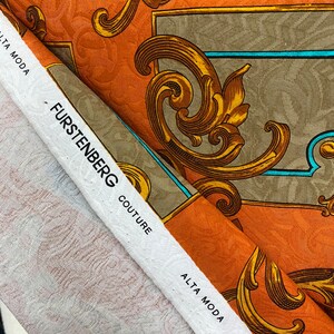 K&ouml;nnte beinhalten: Nahaufnahme eines Stoffmusters mit einem goldenen und braunen Barockmuster. Der Stoff ist mit "Furstenberg Couture" und "Alta Moda" beschriftet.
