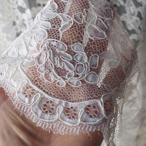 May include: Close-up of delicate white lace fabric. The lace features a floral pattern with intricate details and a scalloped edge. The fabric is semi-sheer, allowing light to pass through, highlighting the detailed design.