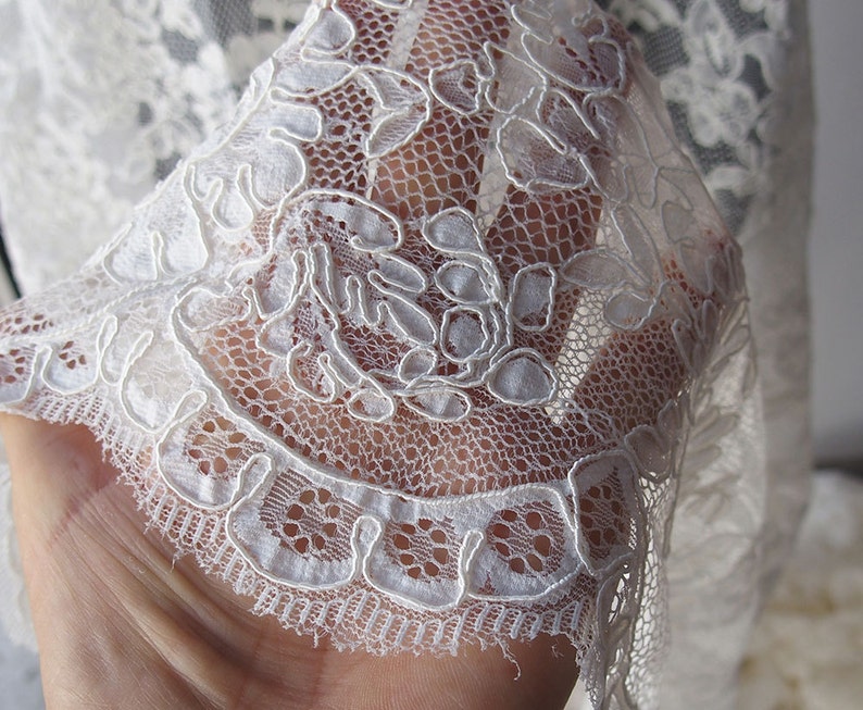 May include: Close-up of delicate white lace fabric. The lace features a floral pattern with intricate details and a scalloped edge. The fabric is semi-sheer, allowing light to pass through, highlighting the detailed design.