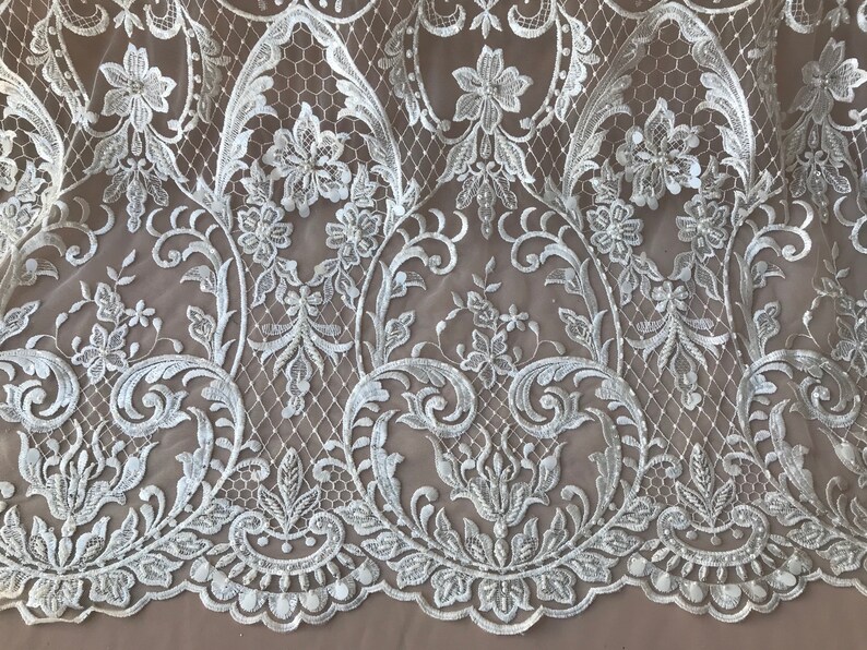May include: White floral lace fabric with a delicate pattern of flowers and leaves. The lace has a scalloped edge and is perfect for adding a touch of elegance to any project.