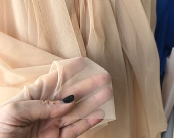 Może przedstawiać: Zbliżenie na przezroczystą, beżową tkaninę o miękkiej, delikatnej fakturze. Tkanina jest drapowana i złożona, tworząc płynny i eteryczny wygląd.