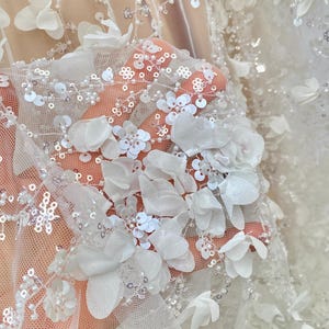 Peut inclure: Tissu de tulle blanc avec des embellissements floraux 3D et des paillettes. Le tissu est transparent et d&eacute;licat, avec une texture douce et fluide.