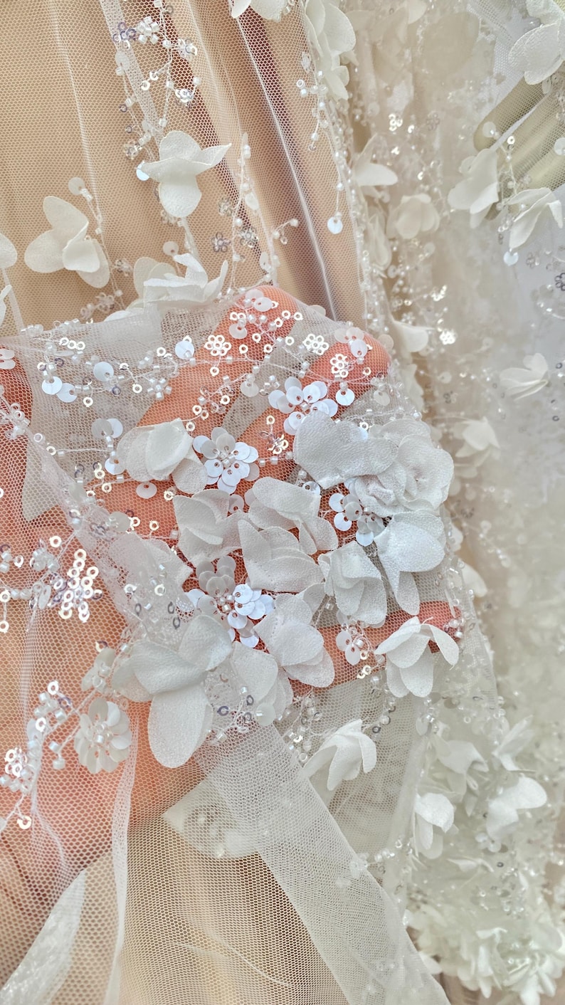 Peut inclure: Tissu de tulle blanc avec des embellissements floraux 3D et des paillettes. Le tissu est transparent et d&eacute;licat, avec une texture douce et fluide.