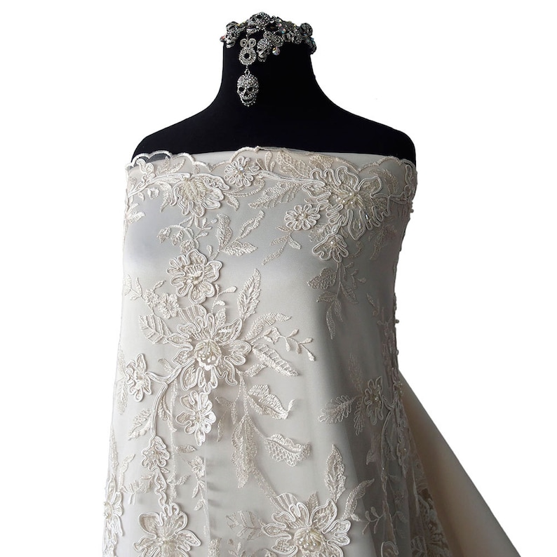 May include: Ivory lace fabric with a floral design and pearl embellishments. The lace is displayed on a dress form.