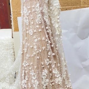 Peut inclure: Un tissu beige transparent avec des embellissements floraux 3D blancs. Le tissu est drap&eacute; sur un mannequin et a un design long et fluide.