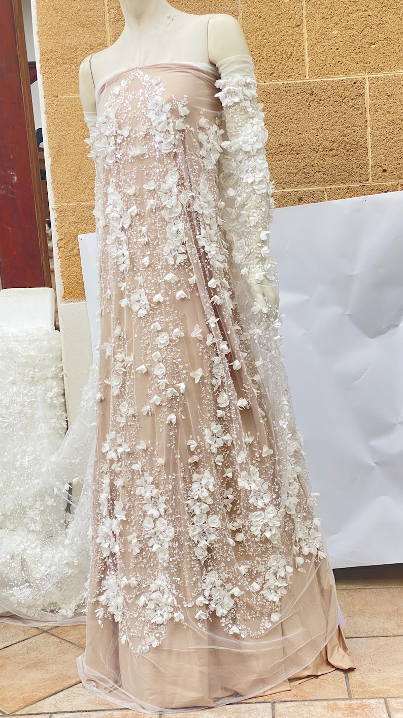 Peut inclure: Un tissu beige transparent avec des embellissements floraux 3D blancs. Le tissu est drap&eacute; sur un mannequin et a un design long et fluide.