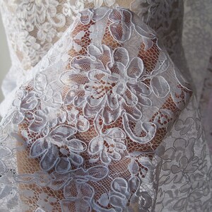 May include: Close-up of white floral lace fabric. The delicate lace features a repeating pattern of large, stylized flowers with intricate details. The fabric is semi-transparent, allowing a glimpse of the background. This lace is suitable for bridal wear, dresses, or other craft projects.