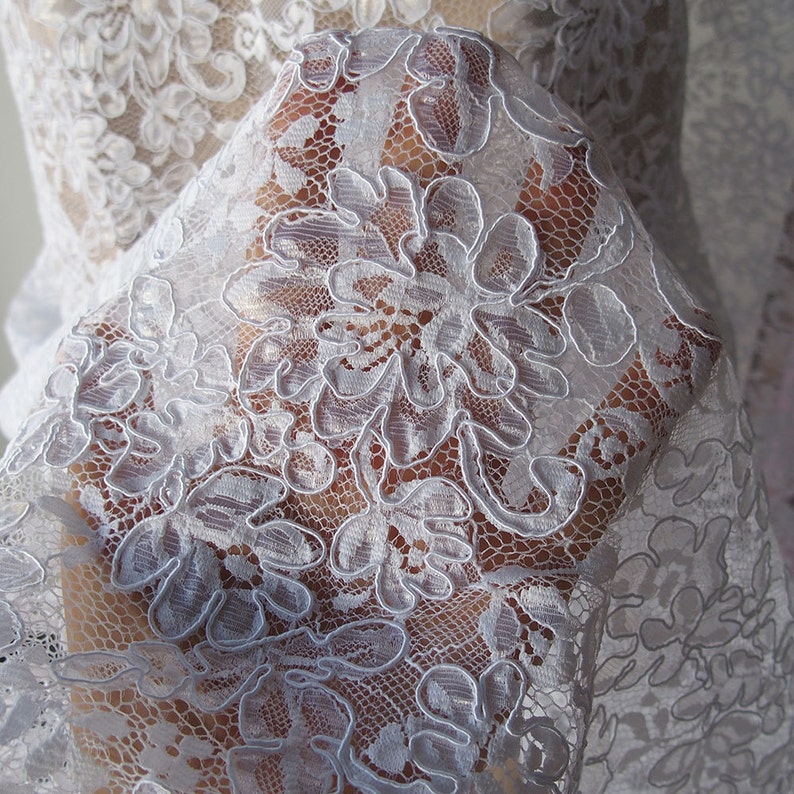 May include: Close-up of white floral lace fabric. The delicate lace features a repeating pattern of large, stylized flowers with intricate details. The fabric is semi-transparent, allowing a glimpse of the background. This lace is suitable for bridal wear, dresses, or other craft projects.