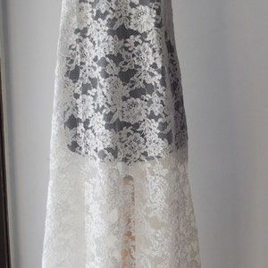 May include: White floral lace fabric draped over a dress form. The lace has a delicate, repeating pattern of flowers and leaves. The fabric is semi-sheer, revealing the form beneath. A decorative necklace is at the top.