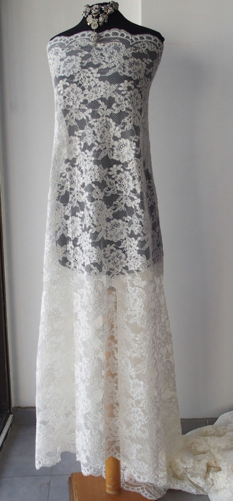 May include: White floral lace fabric draped over a dress form. The lace has a delicate, repeating pattern of flowers and leaves. The fabric is semi-sheer, revealing the form beneath. A decorative necklace is at the top.