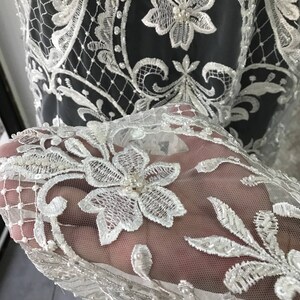 May include: White lace fabric with a delicate floral pattern and pearl embellishments. The lace is sheer and has a soft, flowing texture.
