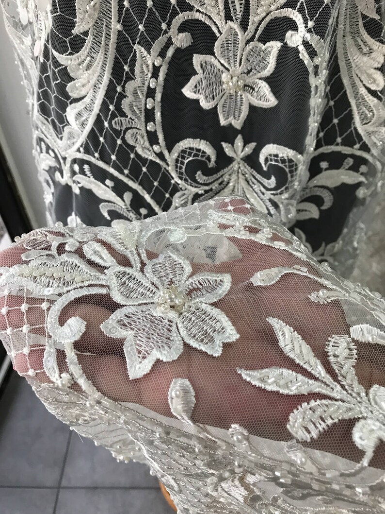 May include: White lace fabric with a delicate floral pattern and pearl embellishments. The lace is sheer and has a soft, flowing texture.