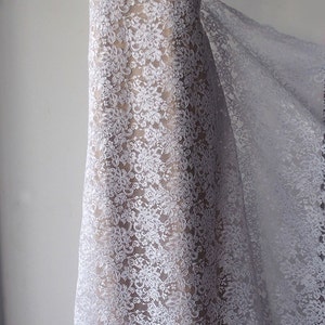 May include: White floral lace fabric draped over a mannequin. The lace features a delicate, repeating floral pattern. A sparkling necklace adorns the mannequin's neck. The fabric is semi-sheer, revealing the form beneath. Suitable for bridal or formal wear.