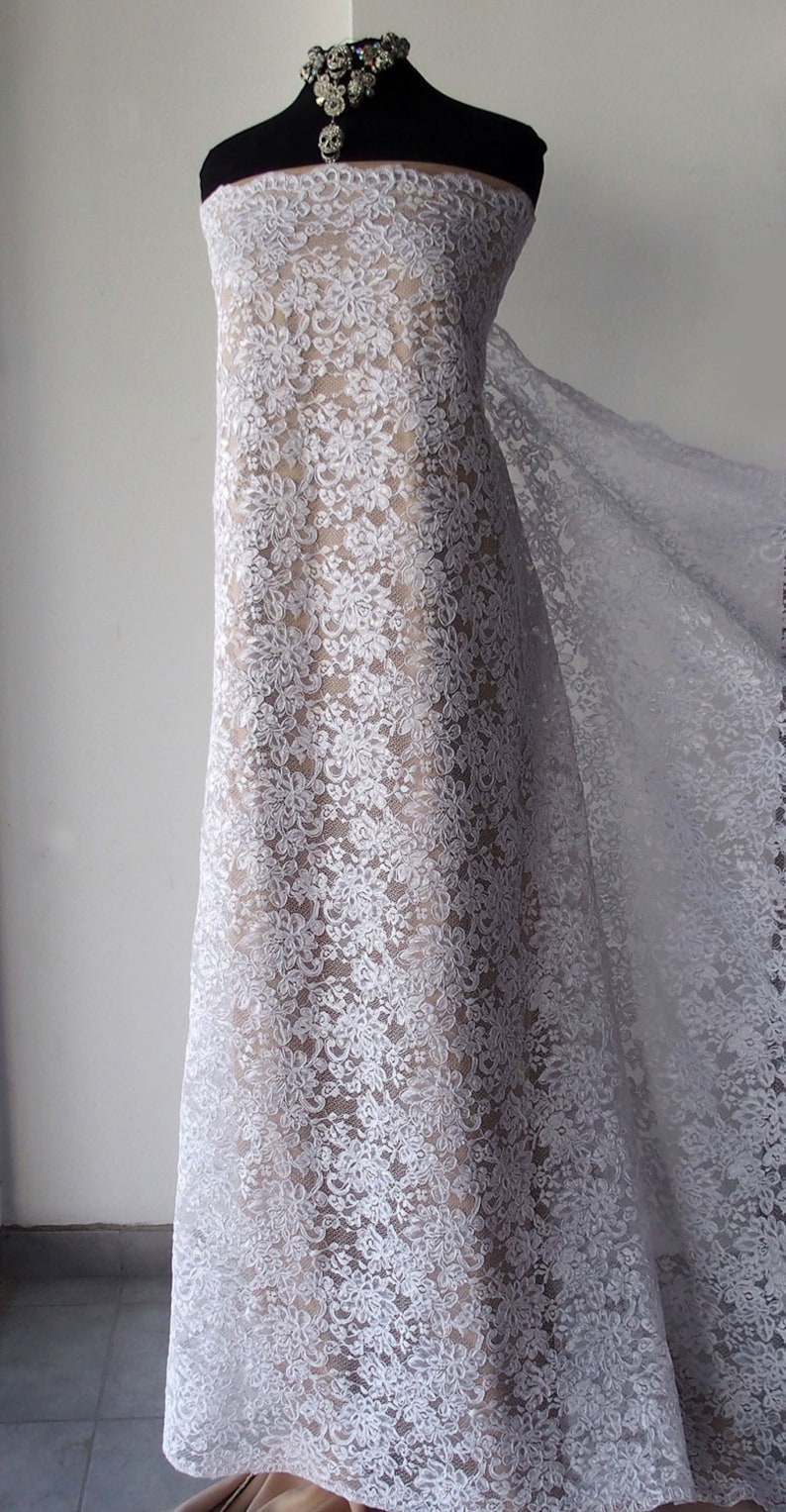May include: White floral lace fabric draped over a mannequin. The lace features a delicate, repeating floral pattern. A sparkling necklace adorns the mannequin's neck. The fabric is semi-sheer, revealing the form beneath. Suitable for bridal or formal wear.