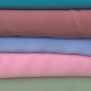 Könnte beinhalten: Ein Stapel Stoff in verschiedenen unifarbenen Farben. Die Farben umfassen Türkis, Mauve, Hellblau, Rosa und Hellgrün. Der Stoff scheint ein weiches, fließendes Material zu sein, möglicherweise Chiffon oder Seide.